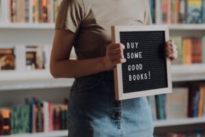 A person holding a board with 'Buy Some Good Books' in a bookstore setting.